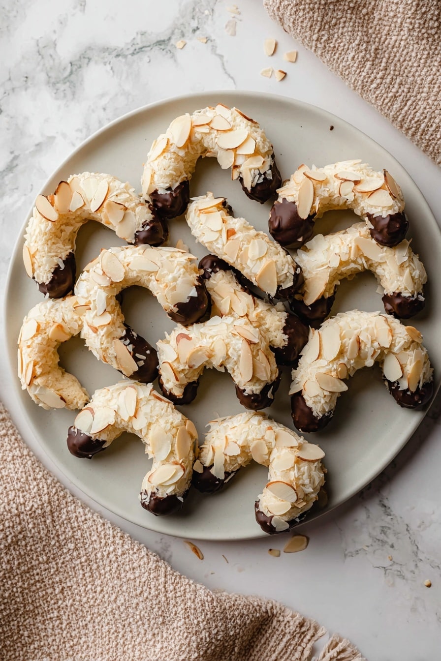 A white round plate holds eleven crescent-shaped cookies arranged in a neat pile. Each cookie has a light beige base, covered with a layer of thin, toasted almond slices that add texture and a warm golden color. The curved ends of each cookie are dipped in dark chocolate, showing a smooth and shiny finish that contrasts with the rough almond surface. The plate rests on a white marbled surface, and a textured beige cloth is partially visible to the right. Photo taken with an iphone --ar 2:3 --v 7 - Almond Horns Cookies with Chocolate Drizzle, almond cookies, chocolate drizzled cookies, nutty almond treats, elegant cookie recipes