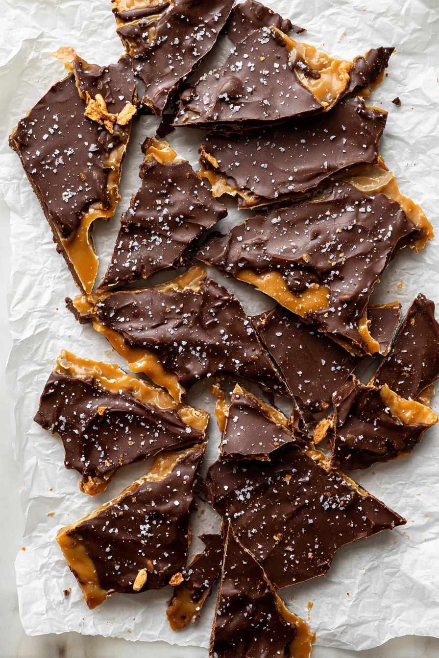 The image shows a broken sheet of chocolate toffee bark spread out on crumpled white parchment paper over a white marbled surface. The bark has two visible layers: a bottom layer of golden, glossy toffee with a smooth texture, and a top layer of dark brown chocolate with a shiny and slightly wavy texture. The chocolate layer is thick, with some small nut pieces and a light sprinkling of coarse sea salt crystals on top, creating contrast. The bark is broken into irregular triangle and shard shapes, scattered closely together, showing rough, jagged edges with some toffee pieces exposed beneath the chocolate. photo taken with an iphone --ar 2:3 --v 7 - Chocolate Pretzel Crack Cookies, sweet salty cookies, easy cookie recipes, peanut butter pretzel cookies, toffee chocolate dessert