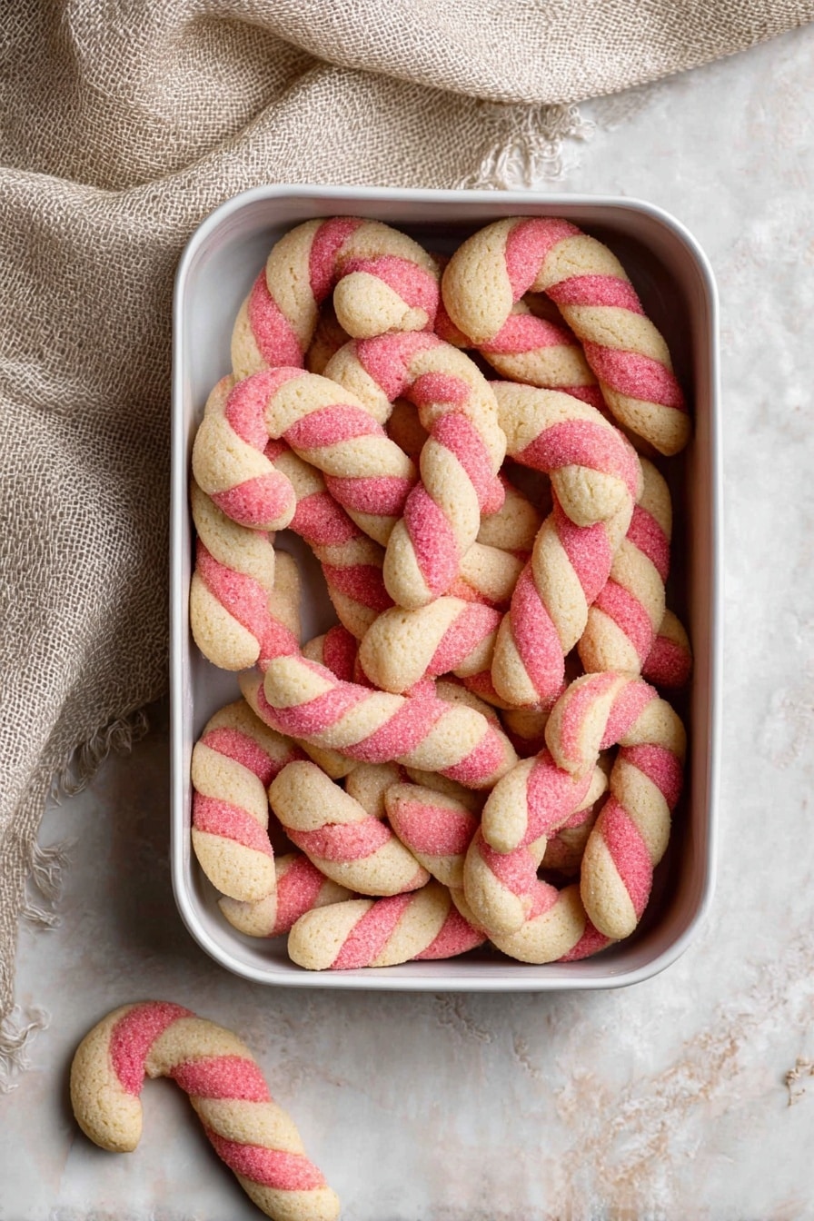 A white round tin full of small candy cane-shaped cookies, each with two twisted stripes—one red and one pale beige—giving a soft, crumbly texture. The cookies fill the tin closely, showing a slight roughness on their surface. One cookie lies outside the tin on the light beige textured surface next to it, and a beige textured cloth is partly draped near the top left corner. The background is a white marbled texture. Photo taken with an iphone --ar 2:3 --v 7 - Candy Cane Cookies with Peppermint Flavor, holiday peppermint cookies, festive peppermint sugar cookies, Christmas peppermint treat, homemade peppermint cookies