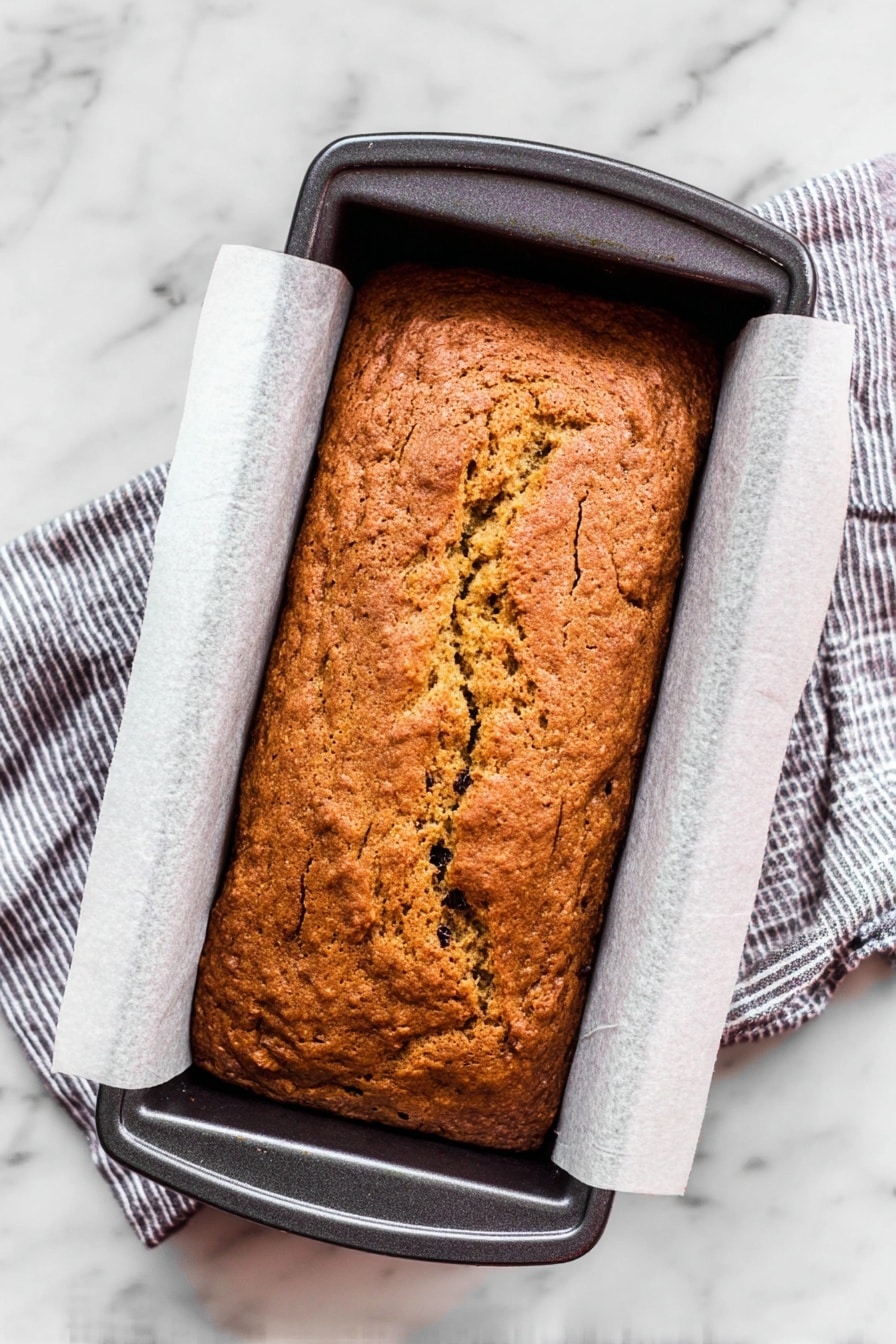 A single rectangular loaf of brown bread with a cracked top surface and visible dark spots is shown inside a black baking pan lined with parchment paper. The bread has a rough texture, and its color varies slightly from golden to deeper brown in some areas, indicating it is baked. The baking pan rests on a striped cloth on a white marbled surface. Photo taken with an iphone --ar 2:3 --v 7 - Yogurt Fruitcake, moist fruitcake with yogurt, dried fruit cake recipe, healthy fruitcake, easy fruitcake dessert