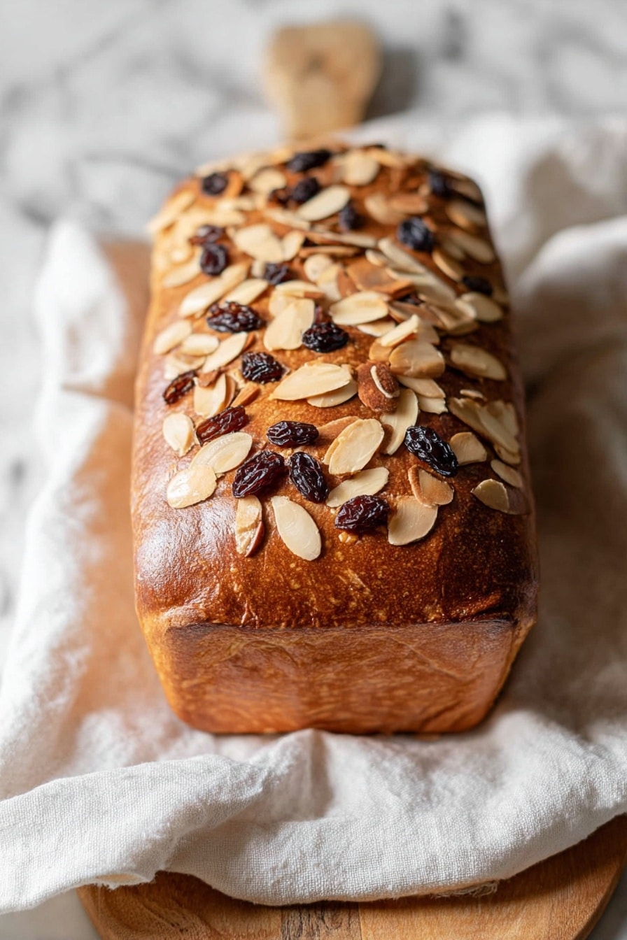 A loaf of raisin bread with a golden brown crust covered in toasted almond slices is placed on a white cloth over a wooden cutting board resting on a white marbled surface. Five thickly sliced pieces of the bread show a soft, light beige interior dotted with dark raisins evenly spread throughout each slice. The bread slices are arranged casually around the loaf, giving a homely and warm feeling. photo taken with an iphone --ar 2:3 --v 7 - Cinnamon Raisin Challah Bread, Challah bread with cinnamon and raisins, homemade cinnamon raisin challah, braided cinnamon raisin bread, soft challah with raisins