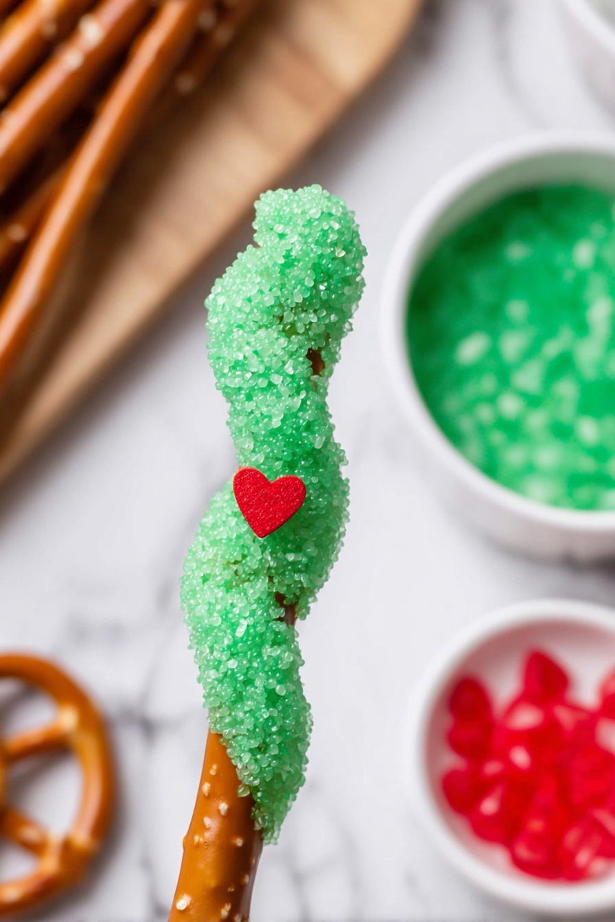 Nine twisted pretzel sticks are lined up vertically on a white marbled surface. Each pretzel stick is coated with a thick green layer of sugar crystals that give a rough, sparkly texture, covering about three-quarters of each stick from the top down. On the green sugar layer of almost every pretzel, there is a small bright red heart-shaped candy placed near the middle or top area. The brown twisted sections of the pretzels are visible at the bottom where the coating ends, showing their shiny and smooth texture. There is a small green pine twig in the upper right corner and a hint of a red object at the top edge of the image. photo taken with an iphone --ar 2:3 --v 7 - Grinch Caramel Pretzel Rods, festive holiday treats, Christmas dessert ideas, green chocolate pretzels, holiday party snacks