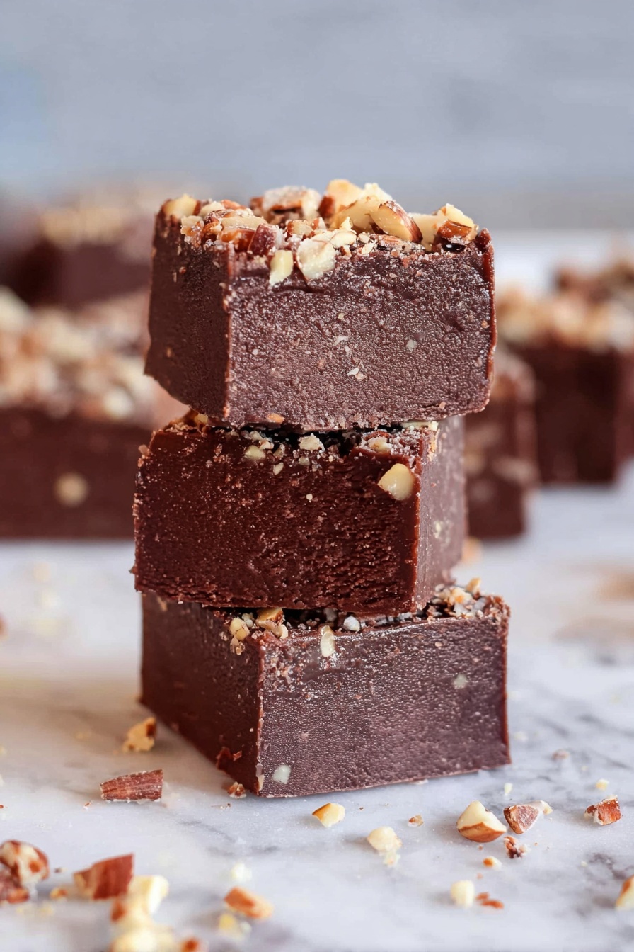 Three thick square pieces of chocolate fudge are stacked vertically, each layer dark brown with a dense and slightly rough texture. The top surface of each fudge piece is sprinkled with small light brown nut pieces, adding a bit of crunch and color contrast. The stack rests on a white marbled surface, with more fudge squares and scattered nut bits blurred in the background, giving a cozy and inviting feel. photo taken with an iphone --ar 2:3 --v 7 - Kahlua Chocolate Peanut Butter Fudge, coffee-infused chocolate fudge, homemade peanut butter fudge, easy alcohol-infused dessert, rich boozy fudge