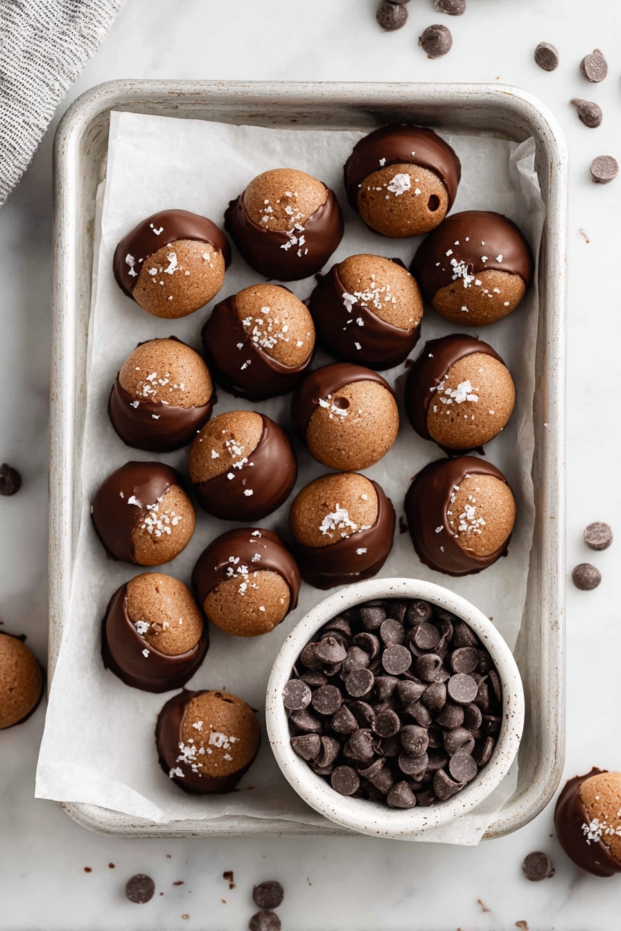 A white tray lined with white parchment paper holds two types of treats: the main layer consists of small round cookies that are dipped halfway in smooth, dark chocolate, with the undipped top part showing a rough matte light brown color, topped with small flakes of white salt. The cookies have slight cracks and a small hole in the center. In one corner of the tray, a small white speckled bowl filled with many dark brown chocolate chips sits on the parchment, surrounded by a few scattered chocolate chips on the white marbled surface around the tray. photo taken with an iphone --ar 2:3 --v 7 - Nutella Buckeye Balls with Chocolate Dipping, Peanut Butter Nutella Bites, Easy Nutella Dessert, Chocolate Dipped Buckeyes, No-Bake Nutella Treats