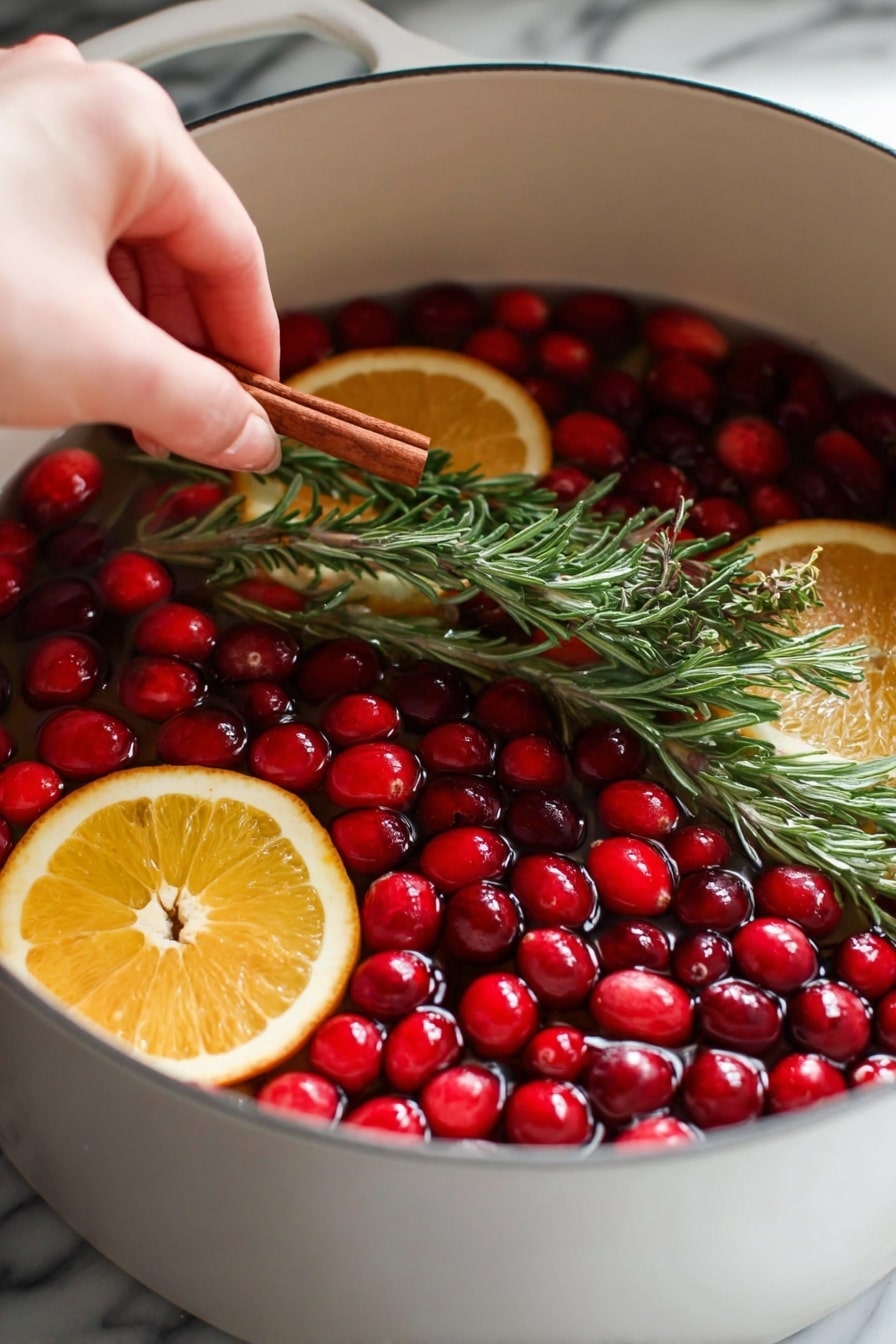 A white pot filled with water holds bright red cranberries mostly floating on top. There are two thinly sliced orange rounds, one near the front and one further back, with a light orange peel around each slice. Several green rosemary sprigs with needle-like leaves float among the cranberries and oranges. A woman's hand is placing a cinnamon stick alongside one rosemary sprig near the center of the pot. The background shows a white marbled texture. photo taken with an iphone --ar 2:3 --v 7 - Festive Christmas Simmer Pot, holiday aroma recipe, Christmas room scent, holiday crockpot recipe, cozy winter simmer pot