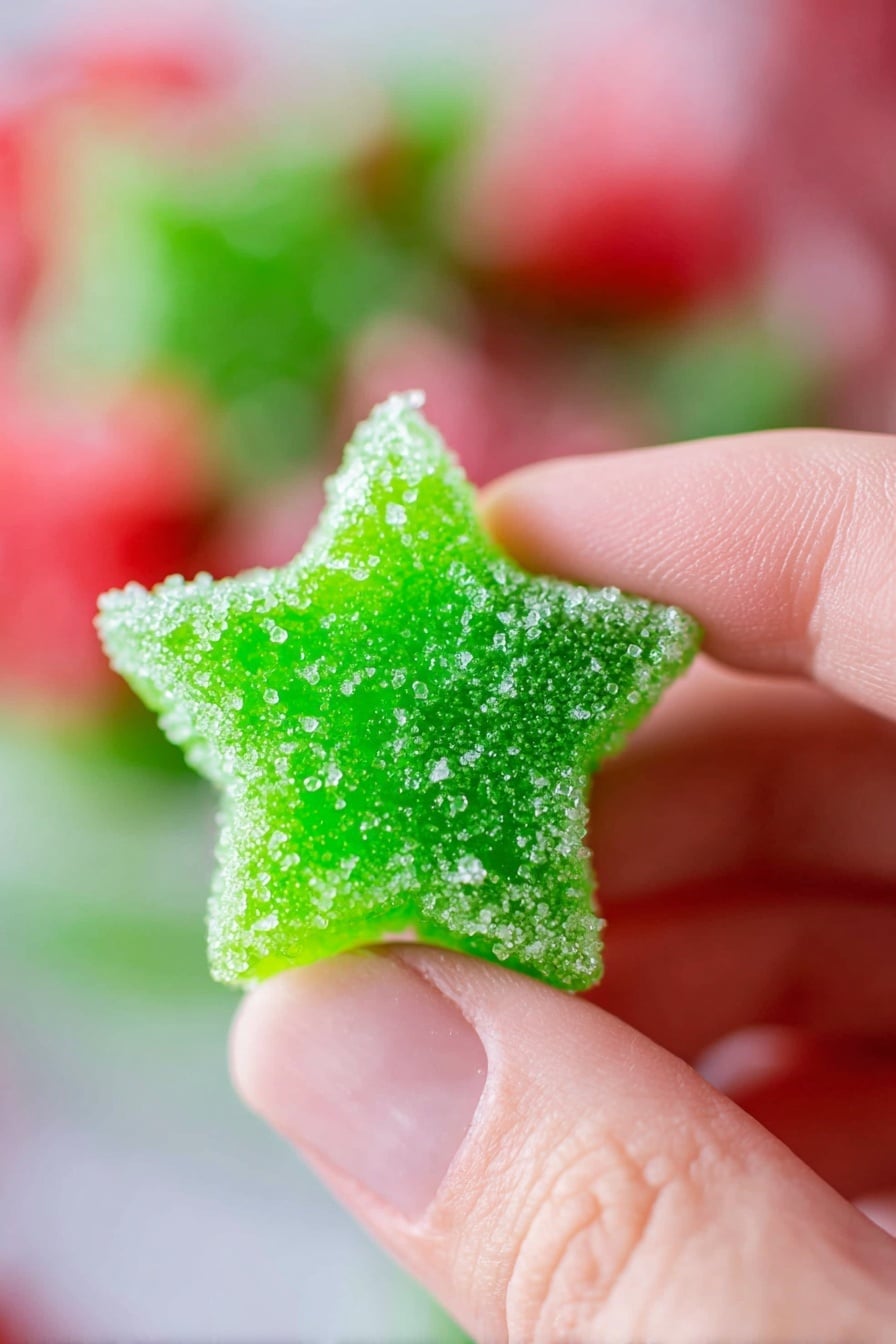 A close-up photo shows a woman's hand holding a bright green star-shaped candy covered in sugar crystals. The candy looks soft and textured, with a rough sugar coat that sparkles under the light. The background is blurred with soft red, green, and white colors suggesting more candies. The overall feel is colorful and festive with the focus fully on the green star candy held gently between the thumb and finger. Photo taken with an iphone --ar 2:3 --v 7 - Homemade Gumdrops, candy making, chewy gumdrop recipe, DIY gumdrops, colorful homemade candies