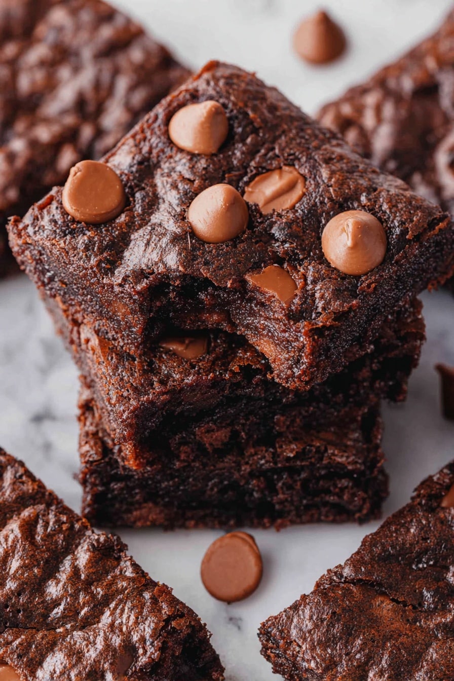 A stack of four thick, dark brown brownies with a rough, textured surface shows melted milk chocolate chips spread through each layer, adding soft spots and shine. The top brownie has a bite taken from the corner, revealing a moist, dense inside. On top of this brownie, there are eight whole, smooth, light brown chocolate chips arranged in a loose circle, with one chip standing upright in the center. The brownies sit on a white marbled surface. Photo taken with an iphone --ar 2:3 --v 7 - Quick Chocolate Cake Mix Brownies, easy chocolate brownie recipe, fudgy brownies with cake mix, simple dessert ideas, quick brownie treats