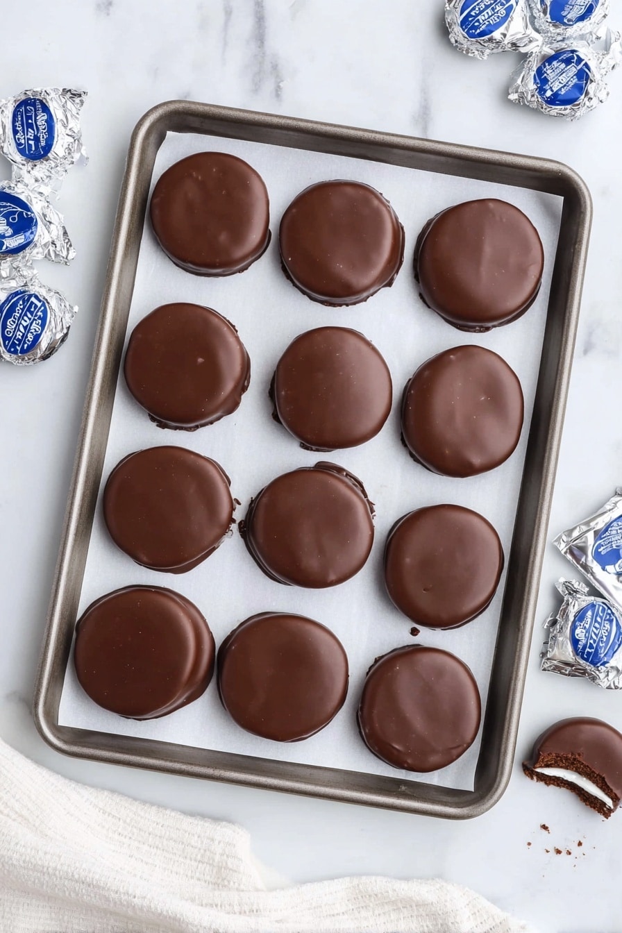 The image shows a baking tray lined with white parchment paper, holding twelve smooth, round chocolate-coated treats arranged in a 3x4 grid. Each piece is thick with a glossy dark brown chocolate layer on top, giving a smooth texture with slight uneven edges. Around the tray on a white marbled surface are silver-wrapped blue and white labeled candies and some unwrapped treats revealing a white creamy center beneath the chocolate coating. One of the treats is partly broken to show the inside. The scene is bright and clean with a white cloth partially visible at the bottom. Photo taken with an iphone --ar 2:3 --v 7 - Homemade Peppermint Patties, peppermint candies recipe, chocolate peppermint treats, DIY peppermint chocolates, easy peppermint candy