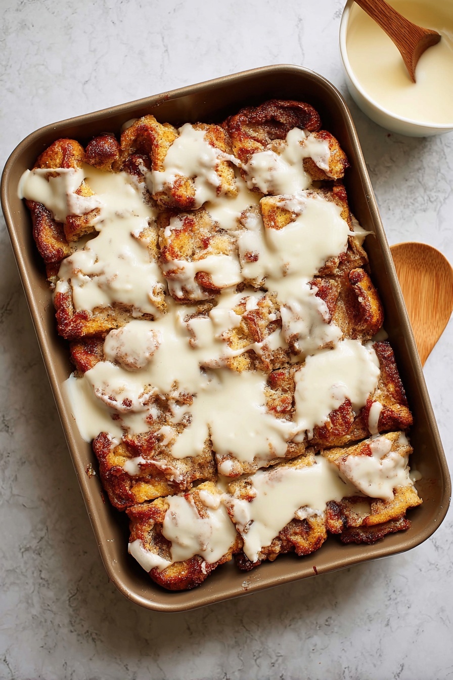 A white plate holds a square piece of cinnamon roll casserole with three clear layers: a light yellow soft base with a slightly browned edge, a thick middle layer of golden brown cinnamon roll chunks, and a creamy white glaze topping that is thick and slightly drizzled over the cinnamon pieces. A silver fork with part of the casserole on it rests on the plate’s edge. In the blurred background, there is another white plate with a similar piece and a white bowl filled with bright red sliced strawberries. The scene is set on a white marbled surface. photo taken with an iphone --ar 2:3 --v 7 - Cinnamon Roll French Toast Bake, easy cinnamon French toast casserole, breakfast bake with cinnamon rolls, make-ahead cinnamon French toast, crowd-pleasing breakfast casserole