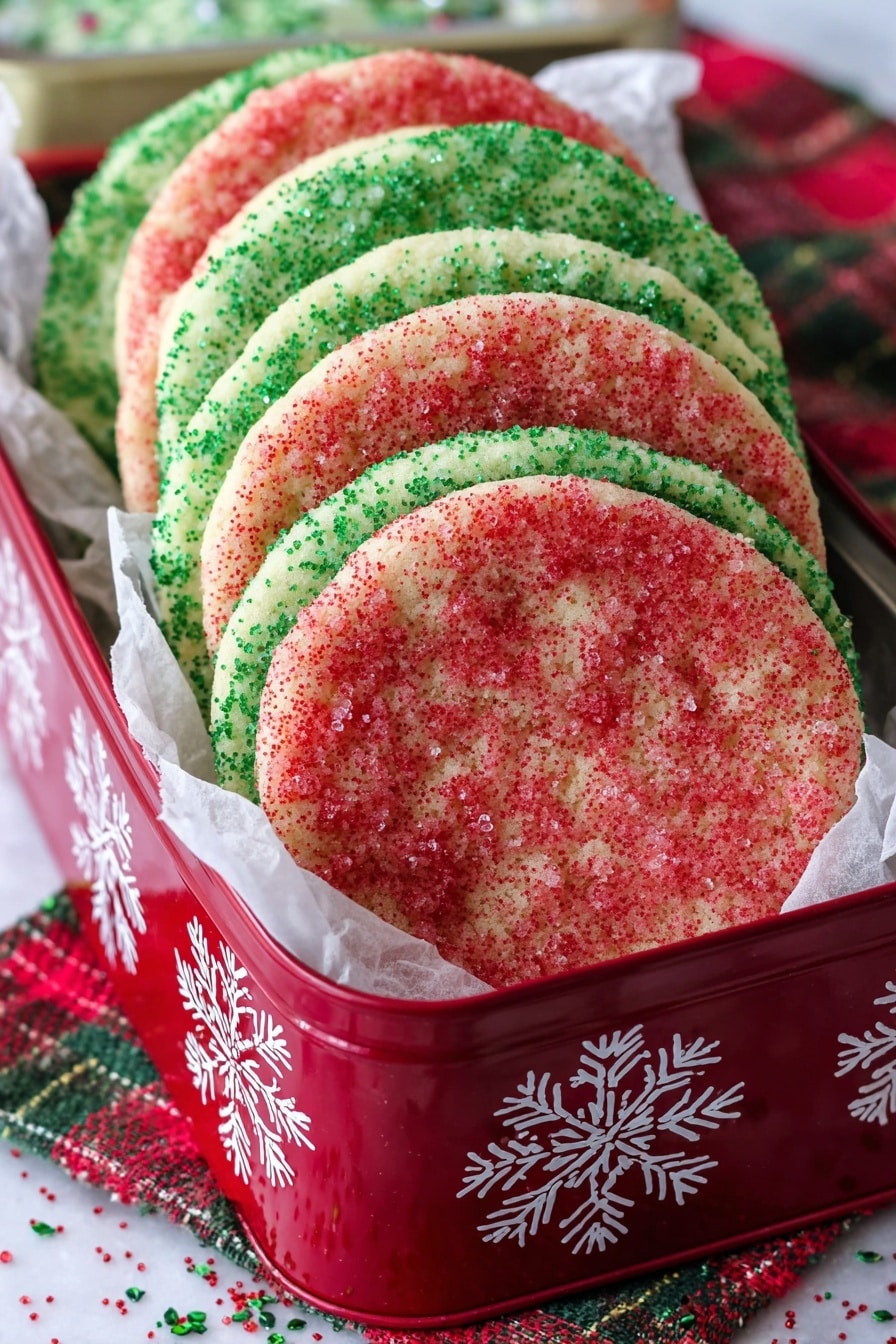 The image shows a stack of large, round sugar cookies arranged vertically inside a red tin box decorated with white snowflakes. There are seven cookies visible, alternating between layers of red sugar sprinkles and green sugar sprinkles. Each cookie has a smooth surface with a slightly raised edge and the sugar crystals give a textured sparkle across the top. The box is lined with white parchment paper that peeks out around the cookies. The tin rests on a white marbled surface with a red, green, and white plaid cloth underneath, with some scattered sprinkles around the base. Photo taken with an iphone --ar 2:3 --v 7 - Christmas Sugar Cookies, Christmas cookies recipe, holiday sugar cookies, festive Christmas cookies, soft sugar cookies for Christmas