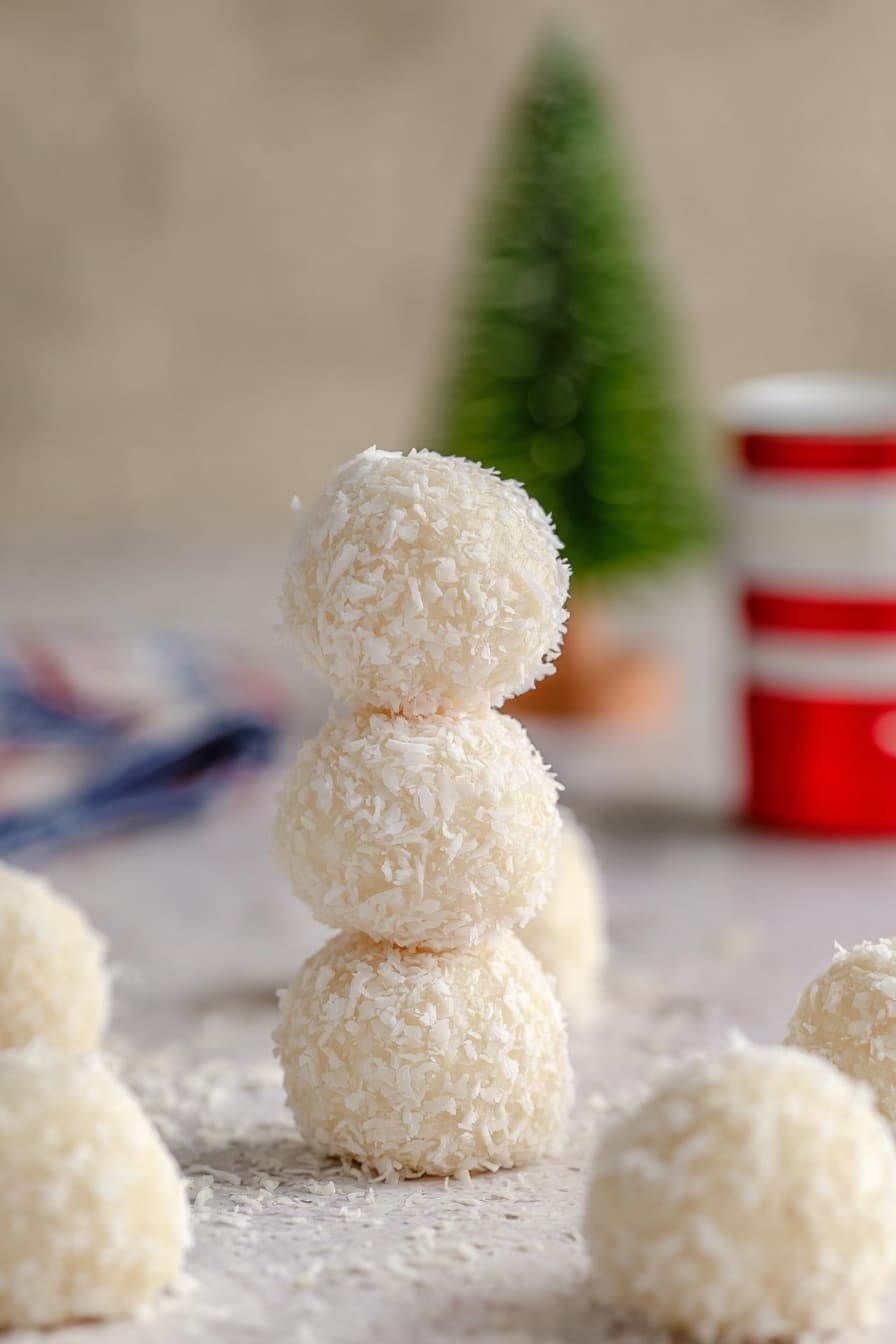 The image shows three white coconut balls stacked vertically in the center. Each ball has a rough texture with small pieces of shredded coconut covering their surface. Around the stack, there are more coconut balls spread out on a light beige surface with a white marbled texture. In the blurry background, there is a small green decoration that looks like a Christmas tree and a red and white cylindrical container on the right. The photo taken with an iphone --ar 2:3 --v 7 - Chocolate Coconut Truffle Balls, Tropical Dessert Treats, Easy No-Bake Truffles, Coconut Dessert Recipes, Party Dessert Ideas