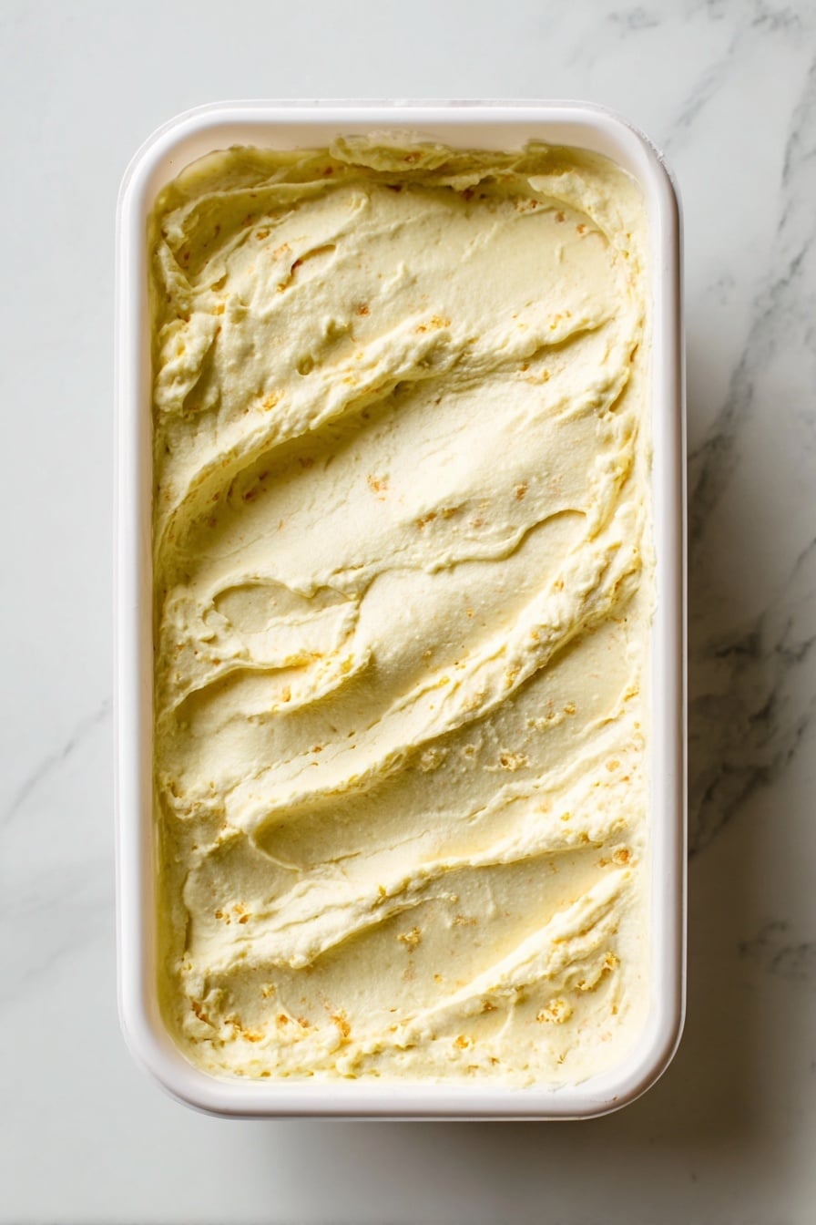 The image shows a white rectangular container filled with a creamy, pale yellow dish that has a slightly uneven, textured surface with small bits of darker yellow or orange mixed in. The top layer appears thick and smooth with a few swirled ridges and small air pockets, indicating a soft, whipped texture. The container rests on a white marbled surface, which adds a subtle contrast to the creamy dish inside. photo taken with an iphone --ar 2:3 --v 7 - No-Churn Banana Pudding Ice Cream, banana pudding ice cream, easy no-churn ice cream recipe, homemade banana ice cream, no-ice-cream maker desserts