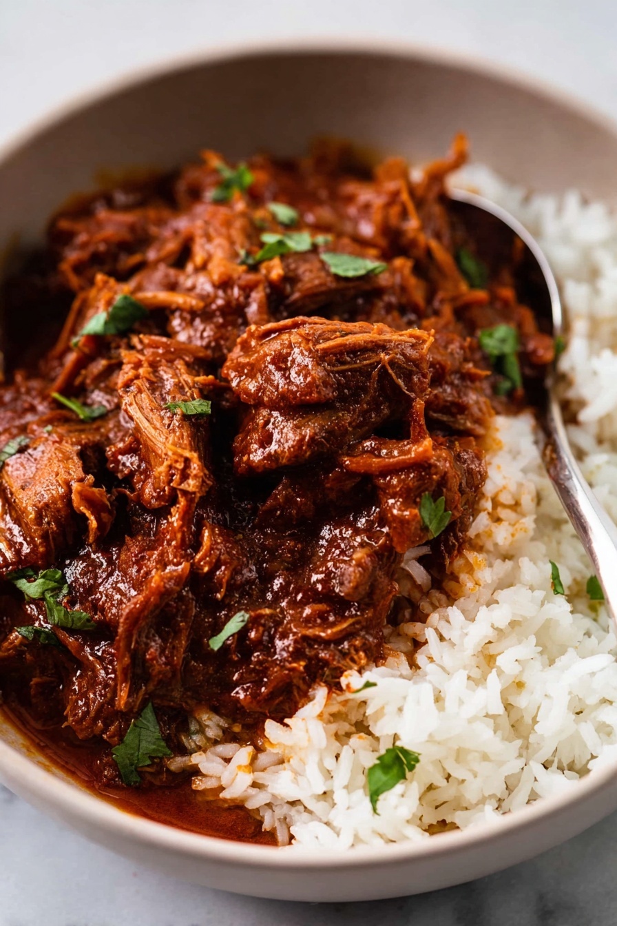 A close-up view of a bowl with two main layers: the bottom layer is white rice with individual grains visible, and the top layer is a rich, dark reddish-brown meat stew with a thick sauce coating tender, shredded meat pieces and larger chunks. The stew is garnished with small green herb leaves scattered on top. There is a silver spoon partially dipped into the rice and stew. The bowl itself is white, set on a white marbled surface. photo taken with an iphone --ar 2:3 --v 7 - Spicy Beef Vindaloo, authentic vindaloo curry, Indian beef curry, spicy curry recipes, beef curry with vinegar