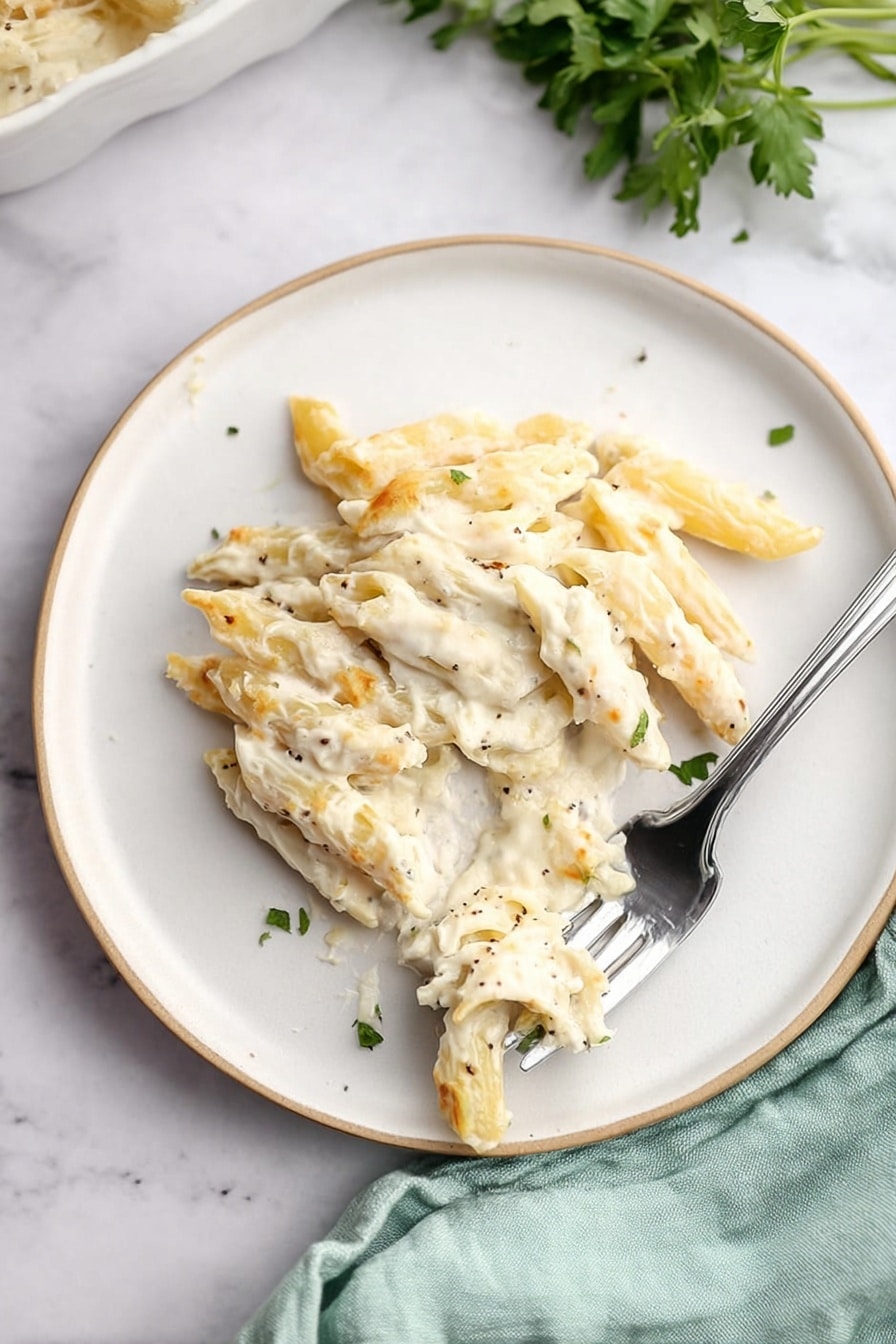 The image shows a close-up of a white spoon holding creamy pasta with a mix of baked penne pasta and pieces of light-colored tender chicken, all covered with melted white cheese that has a golden brown spot on top. Underneath the spoon, a white bowl filled with the same creamy pasta dish is visible, garnished with small green parsley pieces scattered around. The background features a soft light blue cloth and a white marbled surface. photo taken with an iphone --ar 2:3 --v 7 - Quick Chicken Alfredo Pasta Bake, Creamy Pasta Bake, Easy Chicken Pasta Dinner, cheesy casserole with Alfredo sauce, quick weeknight pasta recipe