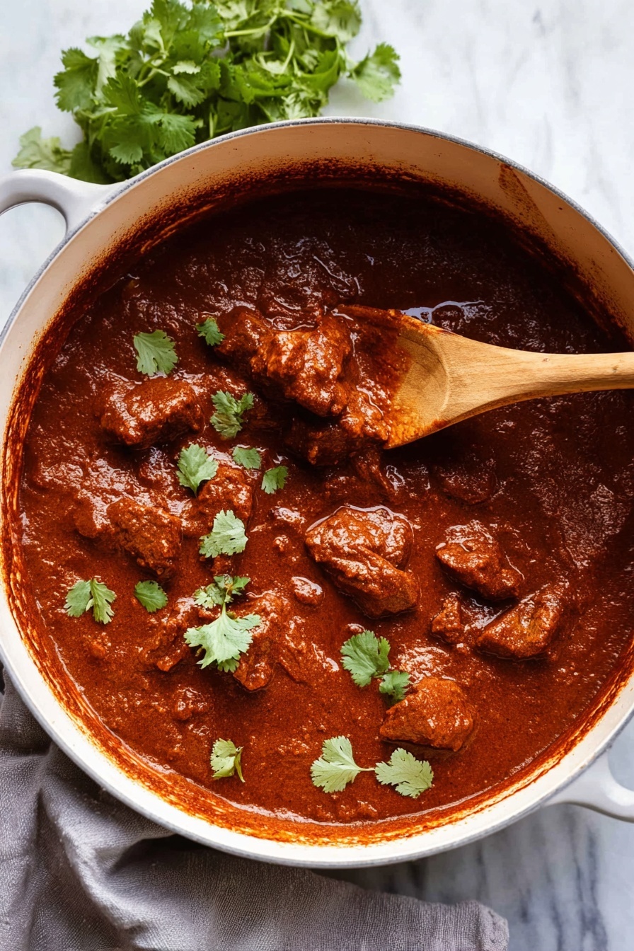 A close-up view of a large white pot filled with thick, rich reddish-brown stew with tender meat chunks evenly spread in the sauce. The stew has a smooth and slightly shiny texture and is topped with fresh green cilantro leaves scattered on top. A wooden spoon is partially submerged on the right side, lifting a piece of meat covered in sauce. The pot sits on a white marbled surface with a small bunch of cilantro in the top left corner and a light gray cloth partially visible beneath the pot. Photo taken with an iphone --ar 2:3 --v 7 - Spicy Beef Vindaloo, authentic vindaloo curry, Indian beef curry, spicy curry recipes, beef curry with vinegar