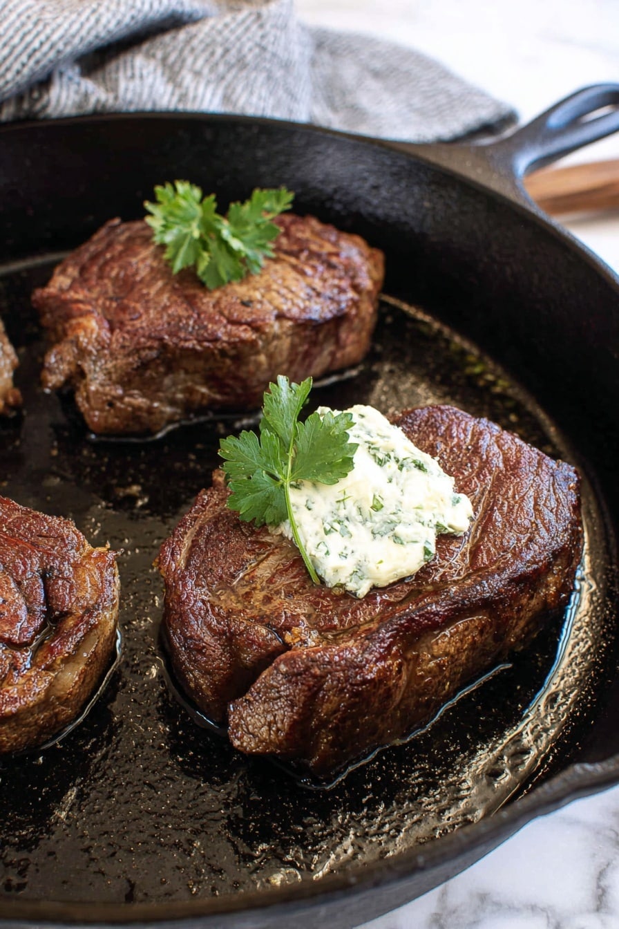 A thick, dark brown cooked steak sits in the center of a white plate, with a round pat of light cream-colored herb butter melting slowly on top. Small green herb leaves are sprinkled on the butter and the steak surface, adding spots of fresh green color. The steak rests in a shallow pool of rich brown juices that shine with a slight oil texture around the base. The white plate is set on a wooden board, and the background shows a blurred light wooden surface with a bunch of fresh green herbs and a gray and white checkered cloth nearby. photo taken with an iphone --ar 2:3 --v 7 - Herb Butter Beef Tenderloin Steak, Beef Tenderloin Steak, Herb Butter Steak, Easy Steak Recipes, Gourmet Beef Dinner