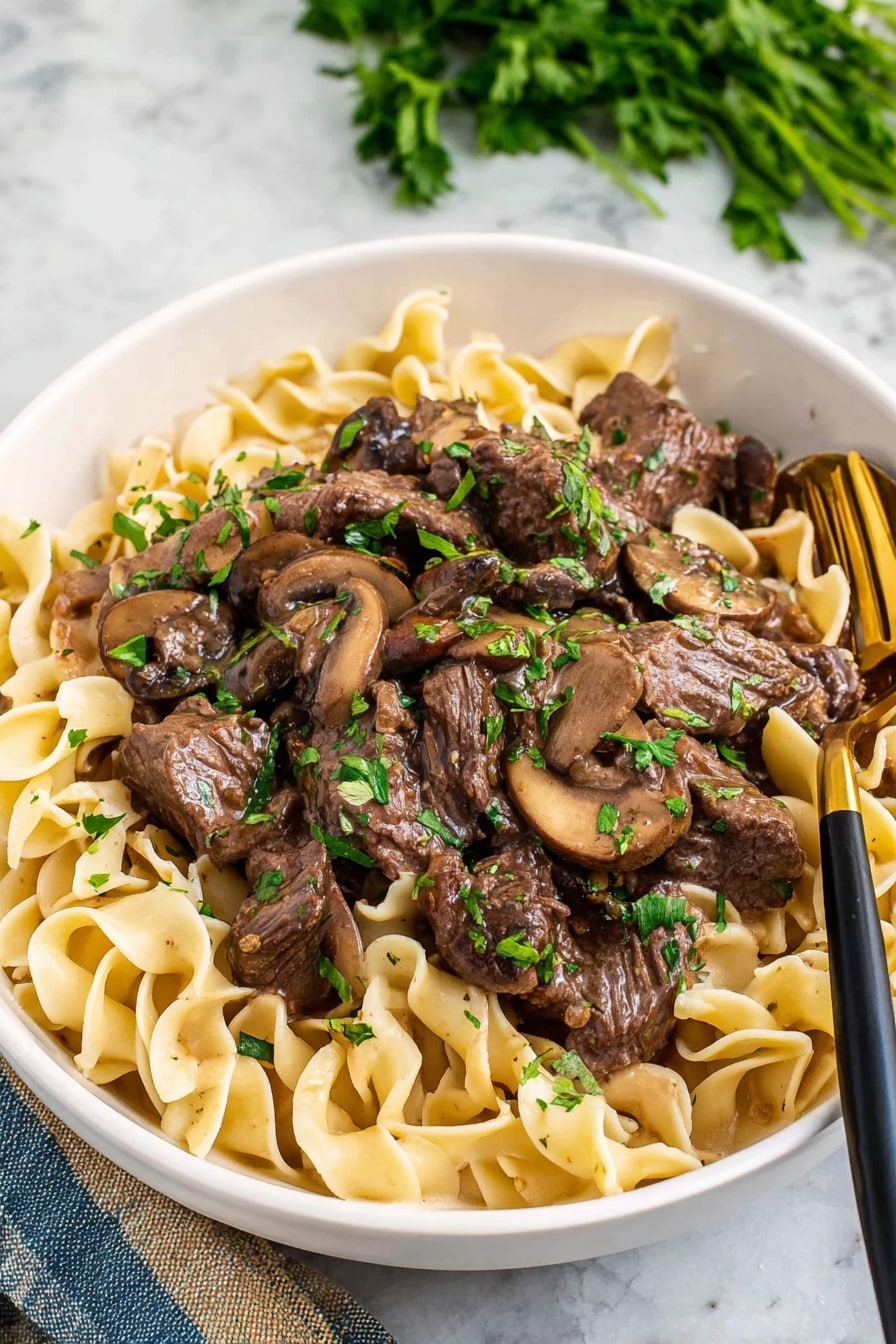 A white bowl filled with rich brown beef stew layered with tender slices of cooked mushrooms, both covered in a glossy brown sauce, scattered with small bright green parsley pieces on top and around the dish, sitting on a white marbled surface with some green herb leaves nearby, photo taken with an iphone --ar 2:3 --v 7 - Crockpot Steak and Mushroom Gravy, easy slow cooker steak recipe, hearty mushroom gravy dinner, tender crockpot beef and mushroom, comfort food steak recipes