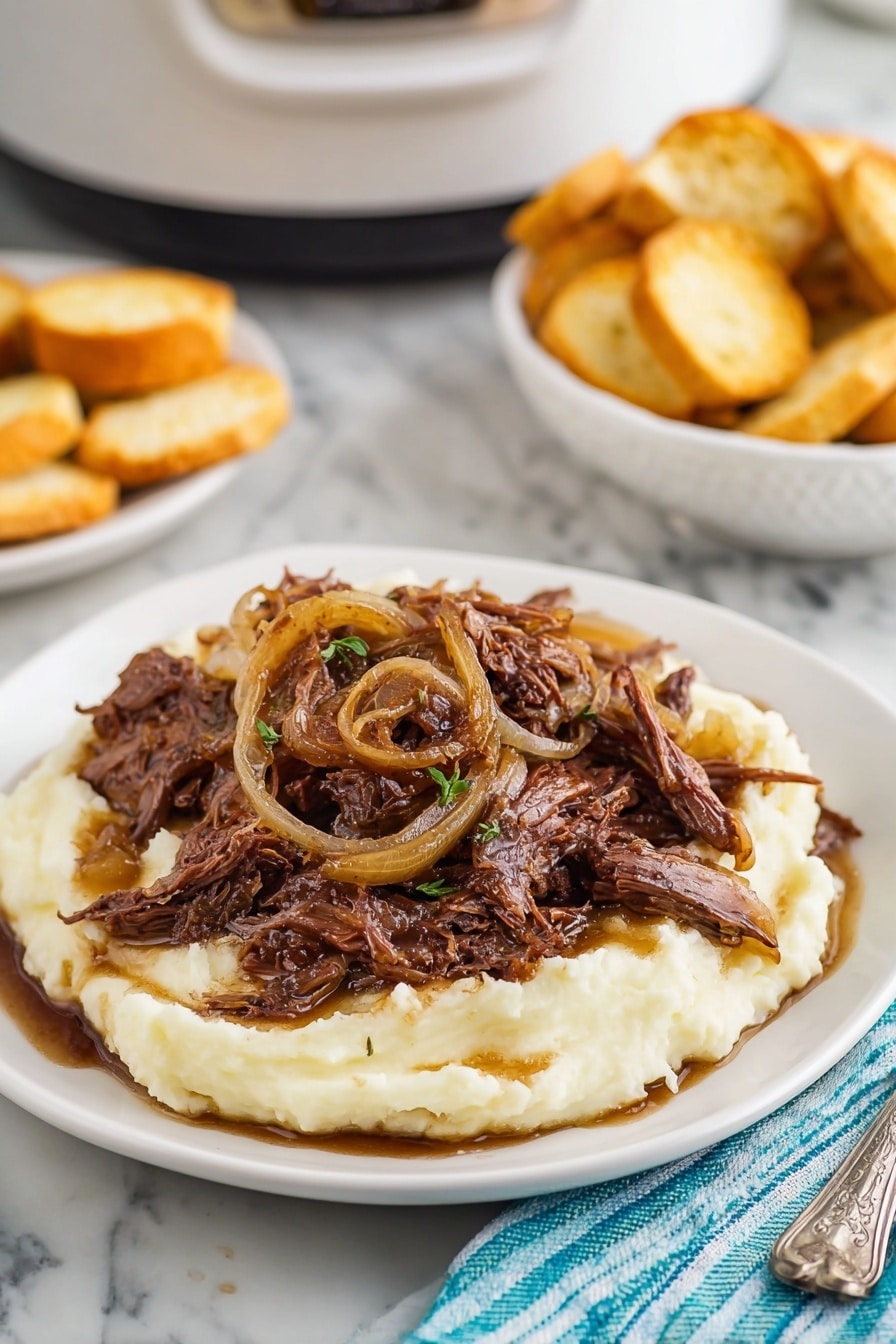 A white plate holds a thick, smooth layer of creamy mashed potatoes as the base. On top is a pile of shredded dark brown meat mixed with soft, caramelized light brown onion slices, some pieces laying loosely and others curled. The meat and onions are glossy with sauce, which slightly drips down onto the mashed potatoes. The plate is set on a white marbled surface, with a silver fork and a blue-striped cloth napkin visible to the right. In the background, a white bowl filled with round, golden brown toasted bread slices and part of a white slow cooker can be seen. Photo taken with an iphone --ar 2:3 --v 7 - Slow Cooker French Onion Roast, French Onion Roast, Easy French Onion Roast, Slow Cooker Roast Recipes, Comforting Dinner Recipes