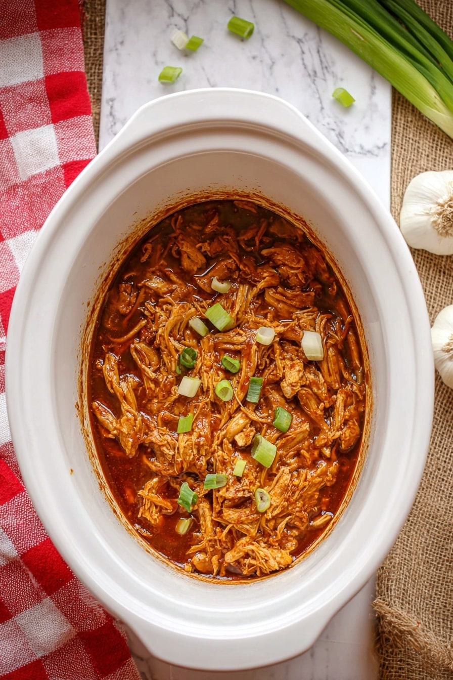 A white slow cooker filled with shredded brownish-orange chicken mixed in a thick dark reddish-brown sauce, garnished with small green onion pieces scattered on top, placed on a white marbled surface. In the background, there are two green onions and a whole garlic bulb on the right side, and a red and white checkered cloth partly visible on the bottom left. Photo taken with an iphone --ar 2:3 --v 7 - Slow Cooker Huli Huli Chicken, Hawaiian chicken recipes, tropical chicken dinner, easy Hawaiian chicken, slow cooker chicken recipes